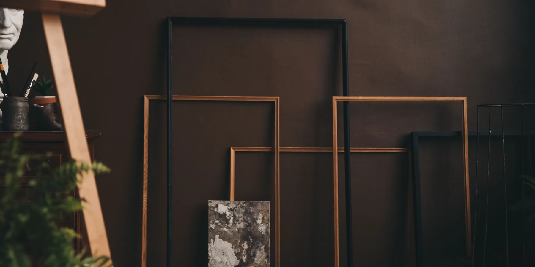 Empty wooden picture frames leaning against a brown wall in a dimly lit art studio with an easel