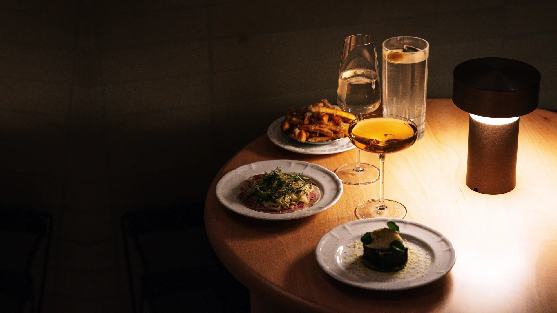A small round table in a dark room, lit by a single lamp and set with plated dishes and a coupe of amber cocktail