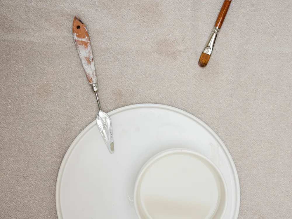 A paint-stained palette knife and a brush lying beside an empty white plate on linen